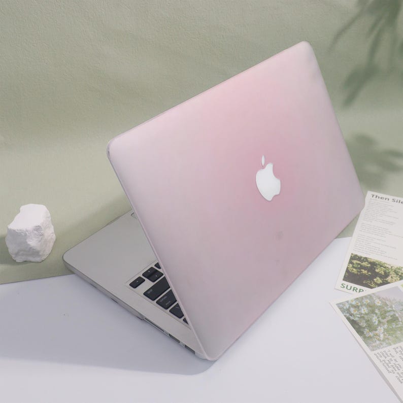 May include: A pink laptop with a white Apple logo on the lid is open on a white surface. The keyboard is visible, and a small white rock and some papers are nearby. The laptop has a silver base.