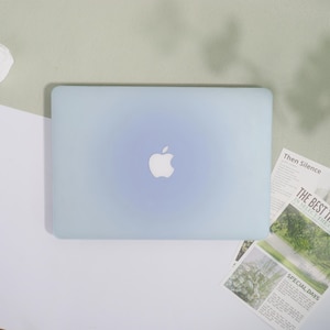 May include: A pale blue laptop featuring the Apple logo. The laptop is placed on a white and green surface. Printed papers with the text "Then Silence", "THE BEST IN SELECT THEA", and "SPECIAL DAYS" are visible.