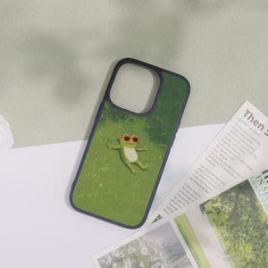 May include: A phone case with a green background featuring a cartoon frog wearing sunglasses, relaxing on grass. The case has a dark blue border and is lying on a white surface.