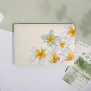 May include: A cream laptop case decorated with a floral pattern of white and yellow plumeria flowers. The flowers are grouped on the right side of the case. The case is rectangular with rounded corners.