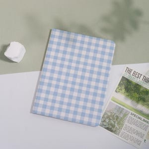 May include: A notebook with a blue and white gingham pattern lies open on a white surface, next to a newspaper clipping. A white, irregularly shaped stone is in the upper left corner. The notebook's cover has a light blue and white checkered pattern.