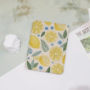 May include: A rectangular notebook with a lemon pattern. The design features yellow lemon slices, whole lemons, green leaves, and small blue flowers on a cream background. The notebook is on a white surface.