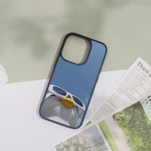 May include: Black phone case featuring a cartoon duck wearing white sunglasses against a blue sky background. The case has a black border and is designed for a smartphone. The duck has a yellow beak.