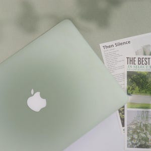 May include: A pale green laptop with a white Apple logo is open on a white surface. Printed materials with the text "Then Silence" and "THE BEST" are next to the laptop. The background is a pale green fabric.