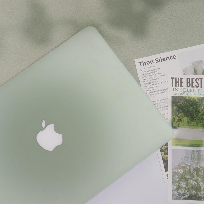 May include: A pale green laptop with a white Apple logo is open on a white surface. Printed materials with the text "Then Silence" and "THE BEST" are next to the laptop. The background is a pale green fabric.