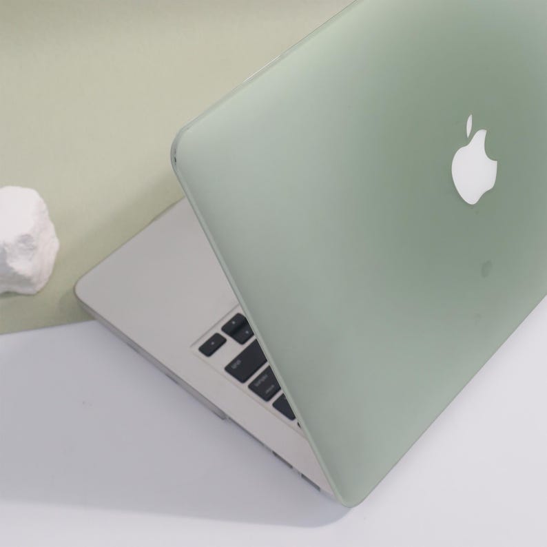 May include: A silver laptop with a pale green cover, displaying the Apple logo. The keyboard is visible, and the laptop is partially open. The background is a soft green and white.