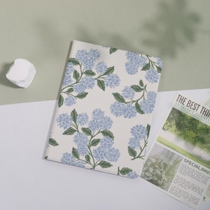 May include: A notebook with a floral design featuring light blue hydrangea flowers and green leaves on a cream background. The notebook is lying on a white surface next to a newspaper clipping.