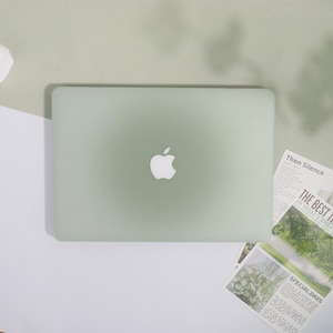 May include: A pale green laptop featuring the Apple logo is positioned centrally. It rests on a light green surface, contrasted by a white and light green backdrop. Newspaper cuttings with the text "Then Silence", "THE BEST THE", and "SPECIAL DAYS" are also visible.
