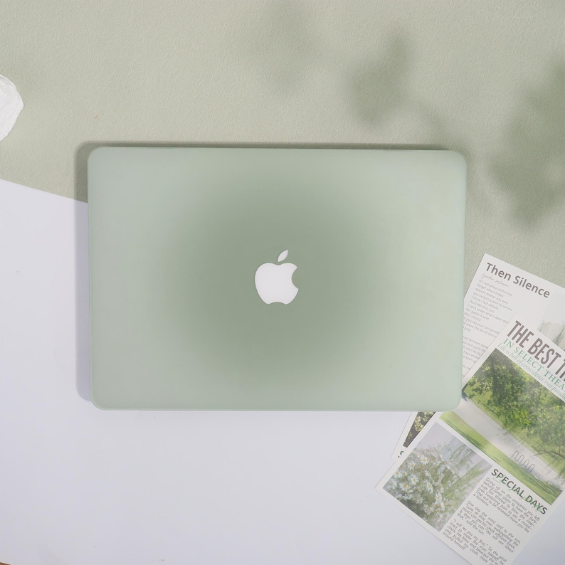 May include: A pale green laptop featuring the Apple logo is positioned centrally. It rests on a light green surface, contrasted by a white and light green backdrop. Newspaper cuttings with the text "Then Silence", "THE BEST THE", and "SPECIAL DAYS" are also visible.