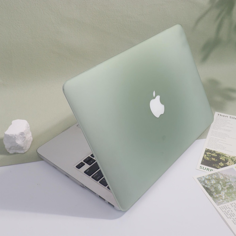 May include: A pale green laptop with a white Apple logo is open on a white surface. The keyboard is visible. A small white rock and some printed papers are next to the laptop.