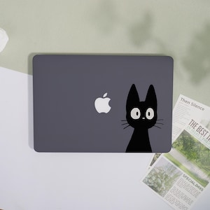 May include: A dark grey laptop featuring a white Apple logo and a black cat design. The cat has large, round eyes and whiskers. The laptop is on a light-coloured surface.
