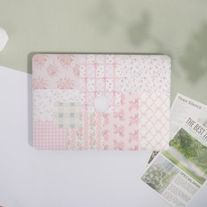 May include: A pink and white floral patterned laptop case. The case has a patchwork design with various floral and bow patterns. The Apple logo is visible in the centre. The background is a light green and white surface.