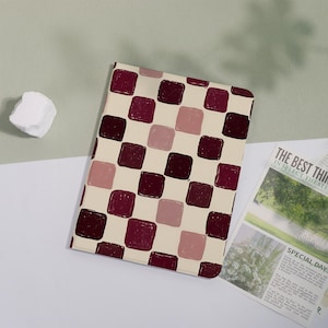 May include: A rectangular notebook featuring a repeating pattern of rounded squares in shades of deep burgundy and muted mauve set against a cream-colored background. The cover has a smooth finish and a consistent grid-like arrangement of shapes.