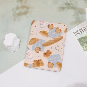 May include: A light beige tablet case featuring a bakery-themed design. The case is adorned with illustrations of croissants, bread, milk bottles, and heart-shaped pastries. The text "gari kory" and "conga bakery" are also visible.