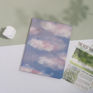 May include: A notebook with a sky-themed cover featuring a blue background with pink and white clouds. The notebook has a glossy finish and a rectangular shape. The image also includes a white rock and a newspaper.