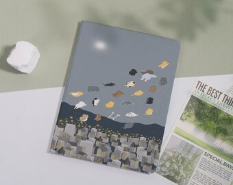 May include: A gray notebook with a whimsical illustration of cats floating above a cityscape. The cover features a night sky design with various cat illustrations in shades of gray, tan, and white. The bottom of the cover shows a city skyline with lit windows.