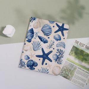 May include: A rectangular notebook featuring a cream-colored cover with a repeating pattern of dark blue seashells, starfish, and coral illustrations. The design includes various shell shapes, including fan-like scallops and spiral conchs, arranged across the surface.