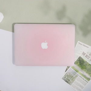 Puede incluir: Un portátil rosa claro con el logotipo de Apple centrado en la tapa. El portátil está sobre una superficie blanca y verde claro. Se ven recortes de periódicos con el texto "Then Silence", "The Best In Select Thea" y "Special Days".