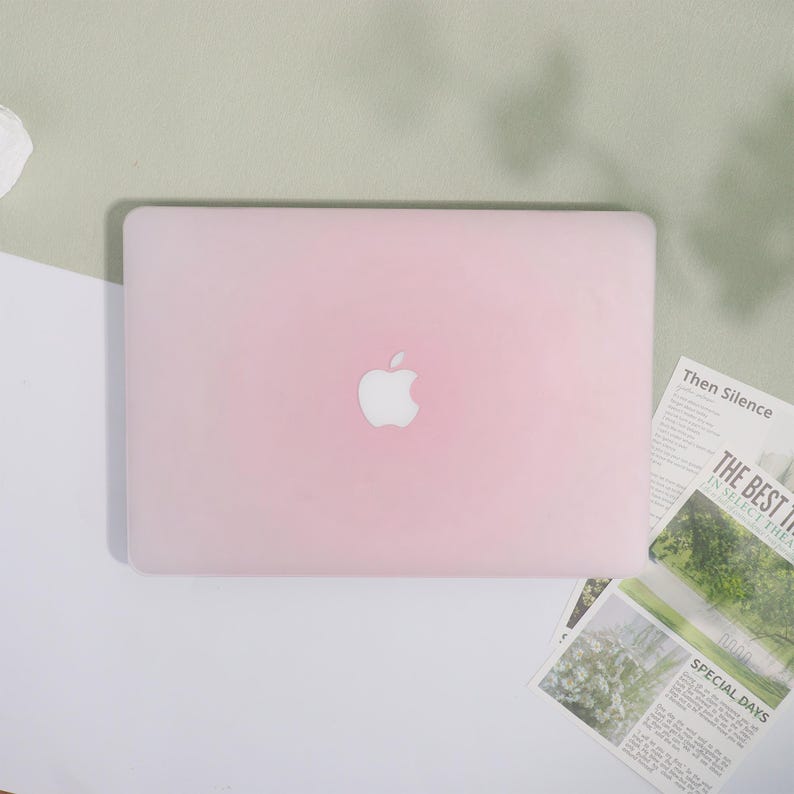 May include: A light pink laptop with the Apple logo centered on the lid. The laptop is on a white and light green surface. Newspaper clippings with the text "Then Silence", "The Best In Select Thea", and "Special Days" are visible.