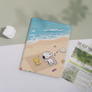 May include: A rectangular notebook cover featuring a beach scene with Snoopy and Woodstock sleeping on a tan towel. The illustration includes blue ocean waves, white foam, a seashell, a starfish, and small colorful pebbles on the sand.