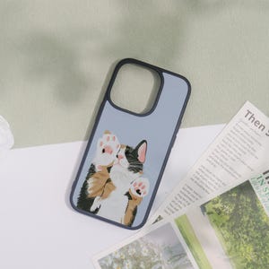 May include: A light blue phone case with a black border. The case features a whimsical illustration of a calico cat with its paws up. The cat has black, brown, and white fur, with pink paw pads.