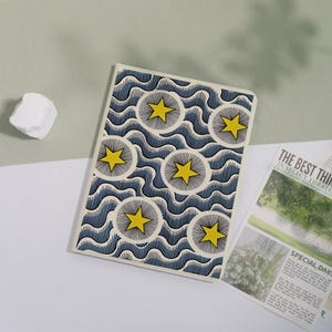 May include: A rectangular notebook featuring a repeating pattern of yellow stars centered within circular sunburst designs, set against a background of dark blue wavy lines. The cover has a cream-colored border and a smooth, matte finish.