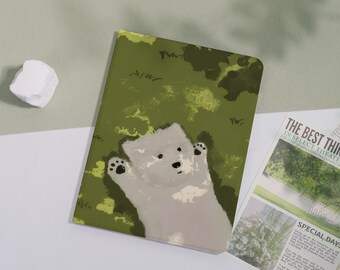 May include: A notebook with a green cover featuring a light gray bear illustration. The bear is lying on its back with its paws up. The background has a green and brown camouflage pattern. A newspaper is visible in the background.