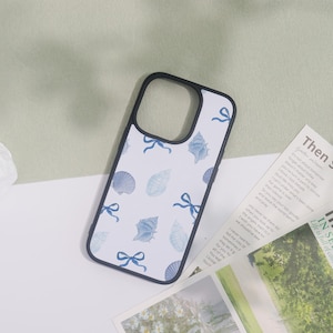 May include: A white phone case with a black border, featuring a pattern of blue seashells and bows. The design is repeated across the case. The case is lying on a white surface.