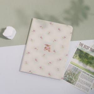 May include: A cream-colored notebook with a floral pattern of small pink roses and a teddy bear illustration. The notebook is lying on a white surface next to a newspaper clipping with the headline "THE BEST THING".