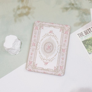 May include: A rectangular, pink and white floral patterned tin box. The design features a central floral medallion and ornate border. The box is on a white and green surface. A white, rock-like object is to the left of the box.