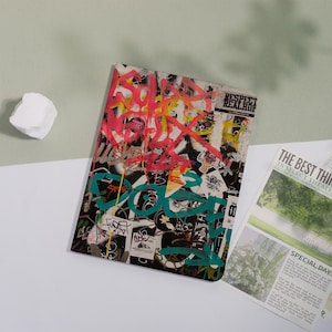 Könnte beinhalten: Ein Notizbuch mit einem Graffiti-Design, das rosa, gelbe und grüne Sprühfarbe auf einem schwarz-weißen Hintergrund aufweist. Das Notizbuch ist mit Aufklebern und Text bedeckt. Eine Zeitung liegt neben dem Notizbuch.