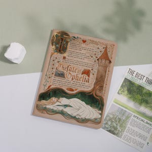 May include: A decorative journal with the title "The Fate of Ophelia" in ornate lettering. The cover features an illustration of a woman in a white dress, a tower, and script. A newspaper with the headline "The Best Thing in Select Theatre" is also visible.
