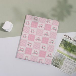 May include: A pink and white patterned notebook featuring a patchwork design with strawberries and bows. The notebook has a soft cover and a grid pattern. A newspaper with the headline "THE BEST THING" is visible in the background.