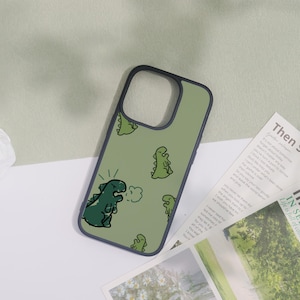 May include: A green phone case with a dark blue border, decorated with cartoon dinosaur illustrations. One dinosaur is shown roaring, with a cloud of breath. The background is a light green colour.