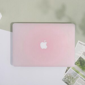 May include: A light pink laptop case with the Apple logo in the center. The case is rectangular and covers the entire top of the laptop. The background is a mix of white and light green.