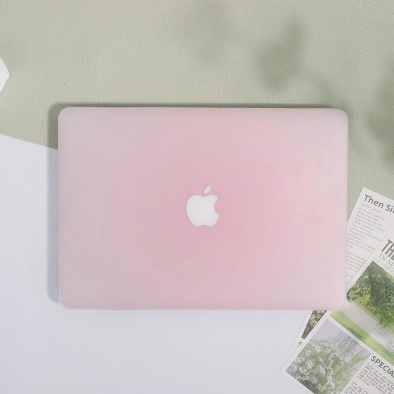 May include: A light pink laptop case with the Apple logo in the center. The case is rectangular and covers the entire top of the laptop. The background is a mix of white and light green.
