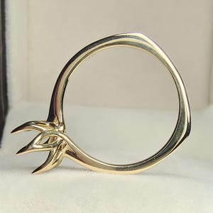 Puede incluir: Un anillo de oro con un diseño único, con una banda circular y un elemento estilizado en forma de flor u hoja. El anillo se muestra dentro de una caja de joyería marrón con interior blanco.