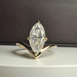 May include: A marquise-cut diamond engagement ring with a gold band. The diamond is set in a prong setting and the band has a V-shaped design. The ring is displayed on a white surface with a dark brown background.