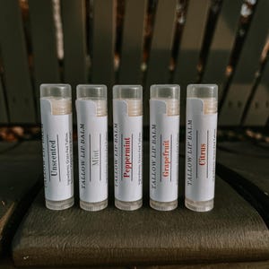May include: Five lip balm tubes with white labels and clear caps. The labels read "Unscented", "Mint", "Peppermint", "Grapefruit", and "Citrus". The lip balms are displayed on a dark wooden surface.