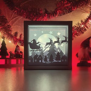 Könnte beinhalten: Quadratische, beleuchtete Weihnachtsdekoration mit einer Silhouette des Weihnachtsmanns in seinem Schlitten, gezogen von Rentieren, über einem verschneiten Dorf und Wald. Das Design ist vor dem Hintergrund eines Vollmonds und Sternen mit einem dunkelgrauen Rahmen.