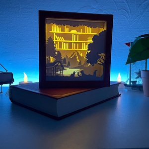 Puede incluir: Una caja de sombras decorativa e iluminada con una escena de bosque, colocada sobre un libro cerrado. La caja presenta una cabaña, árboles y montañas, con un brillo amarillo cálido. Un piano en miniatura y velas están en primer plano.