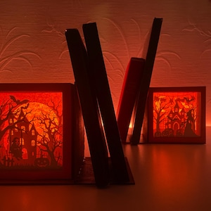 Puede incluir: Soportes para libros rojos con temática de Halloween con un diseño de casa embrujada espeluznante. Los sujetalibros presentan una silueta de una casa embrujada, árboles, murciélagos y una luna llena. La escena está iluminada con un brillo naranja, creando una atmósfera festiva y misteriosa.
