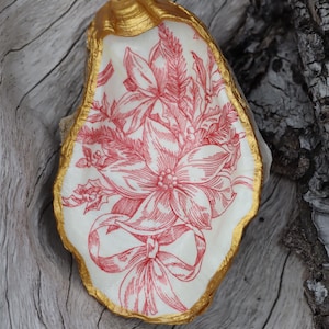May include: A white oyster shell with a gold rim, decorated with a red floral design and a red bow. The shell is sitting on a wooden surface.