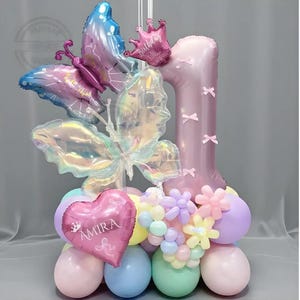 May include: A celebratory balloon display for a first birthday. It includes a large number one balloon, butterfly-shaped balloons, and a heart-shaped balloon with the name "Amira". The arrangement features pastel-coloured balloons.