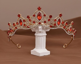 May include: A gold-toned crown tiara with red and clear gemstones. The tiara is displayed on a white pedestal against a brown backdrop. The text "Crown tiara" is at the top of the image.