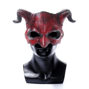Puede incluir: Una máscara de diablo roja con cuernos curvados y aberturas para los ojos. La máscara se muestra sobre una cabeza de maniquí negra sobre un fondo blanco. La máscara tiene un acabado envejecido, lo que sugiere una apariencia antigua.