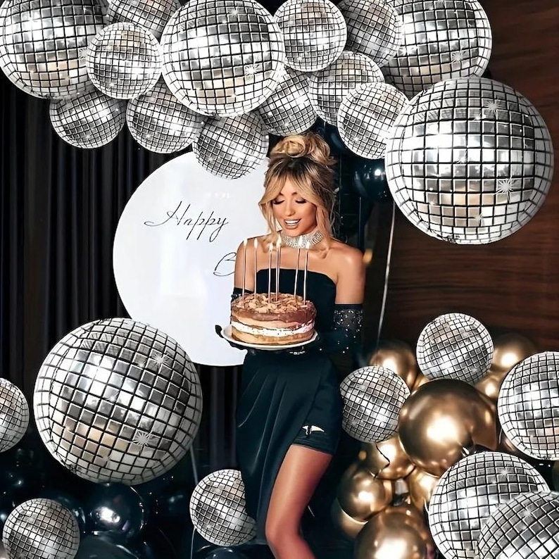 May include: A woman holding a birthday cake with lit candles, surrounded by disco ball balloons and gold balloons. A large white balloon behind her reads "Happy Birthday". The woman is wearing a black dress and a sparkling necklace.