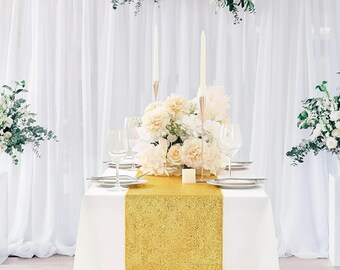 Puede incluir: Una mesa bellamente decorada para un evento especial. La mesa está cubierta con un mantel blanco y un camino de mesa dorado con lentejuelas. El centro de mesa incluye flores blancas, velas y cristalería. Cortinas blancas y arreglos florales completan el look elegante.