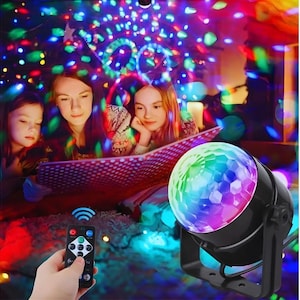 May include: A black disco ball light projector with a colourful, multi-faceted globe. The projector is on, casting a vibrant array of coloured lights. A remote control is held in the foreground. Three people are in the background, illuminated by the lights.