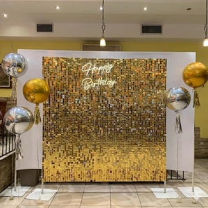 May include: A party backdrop featuring a gold sequin wall with the words "Happy Birthday" in neon script. Silver and gold balloons with tassels are positioned in front of the backdrop, creating a festive atmosphere.